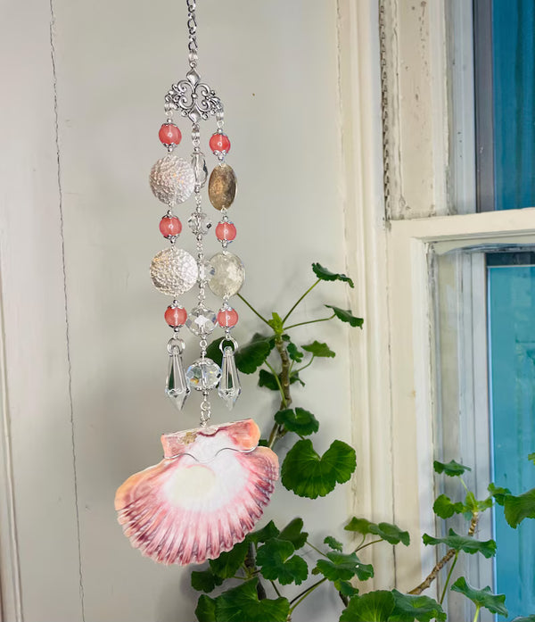 Decorative shell wind chime with beads hanging near a window with plants.
