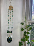 Decorative hanging with green beads and a large green stone in front of a window with a plant.