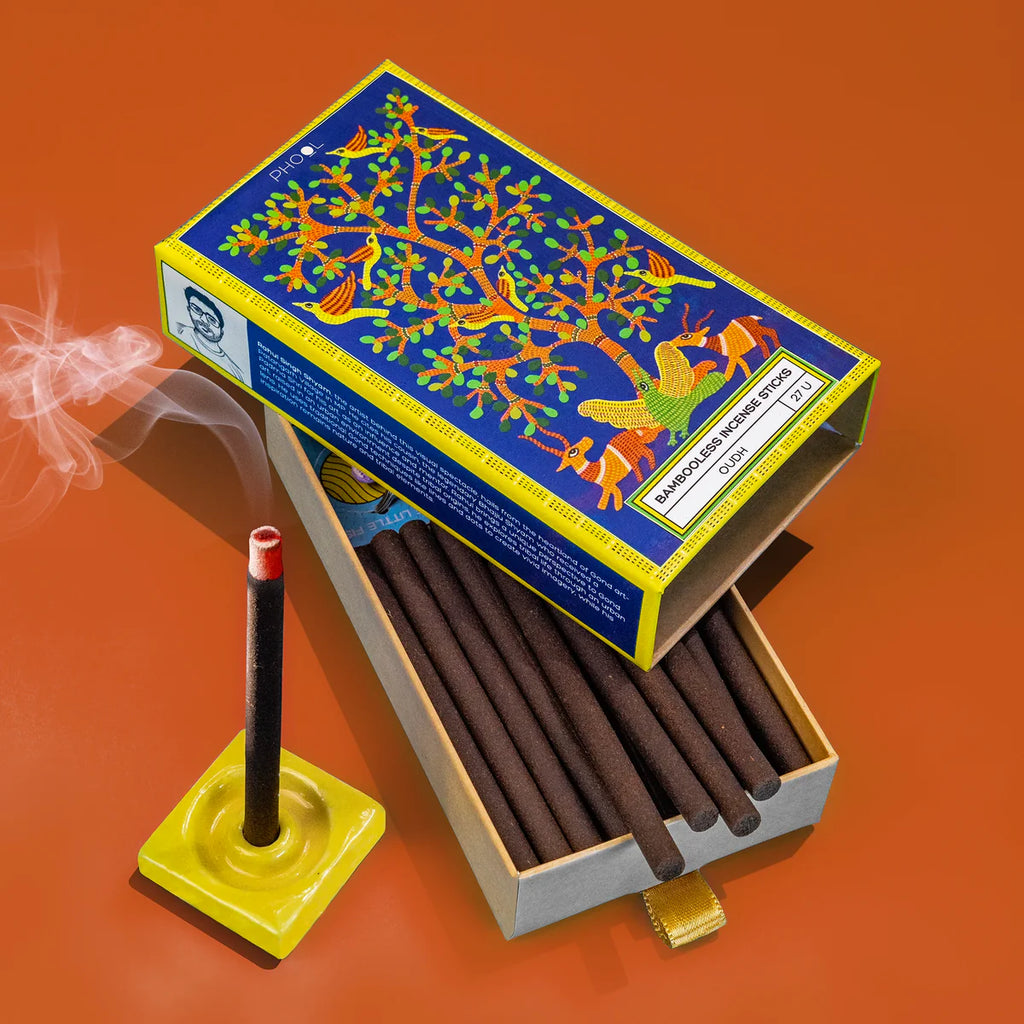 A box of incense sticks with a colorful bird and tree design on the lid, the incense sticks are placed inside a yellow holder on a red background, smoke is rising from one of the sticks indicating it is being burned.