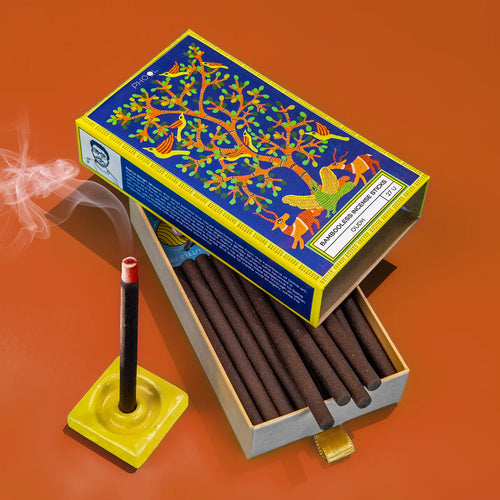 A box of incense sticks with a colorful bird and tree design on the lid, the incense sticks are placed inside a yellow holder on a red background, smoke is rising from one of the sticks indicating it is being burned.
