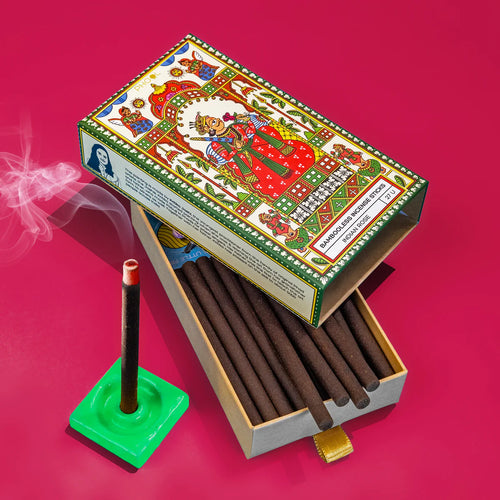 A box of Bambooless Indian Rose Incense with incense sticks inside, against a red background, with smoke rising from one of the sticks, and a decorative lid on top.