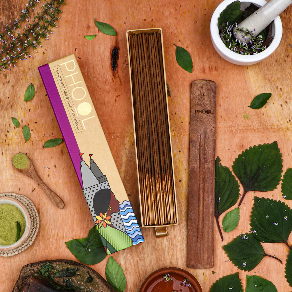 A box of Phool Patchouli Incense Sticks displayed alongside natural ingredients like leaves, spices, and a mortar and pestle, indicating the earthy, musky aroma of the product.