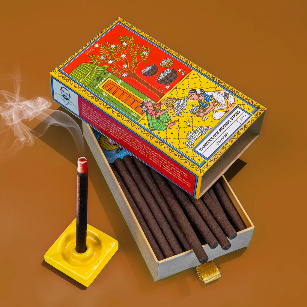 A box of Bambooless Jasmine Incense with incense sticks inside, alongside a yellow ceramic holder and a red lid with decorative illustrations. Smoke is rising from one of the sticks, indicating it is lit.
