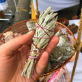 Hand holding a bundle of sage sticks tied with red string, with a blurred background.